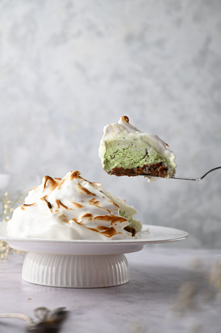 A decadent Baked Alaska slice with a toasted meringue topping, revealing green ice cream and a brown base, is lifted from the whole Baked Alaska on a white cake stand.
