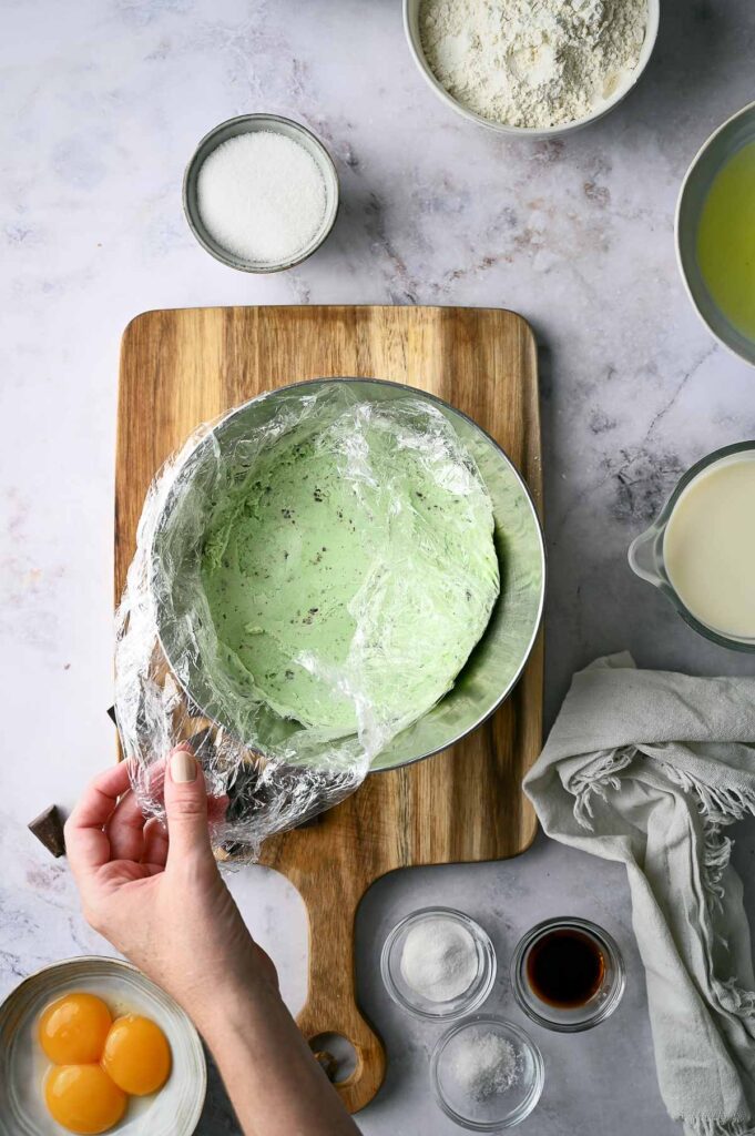 A hand lifts plastic wrap from a bowl of green dough on a wooden board, surrounded by baking ingredients including eggs, sugar, flour, milk, and vanilla—suggesting preparations for an imaginative treat like Baked Alaska.