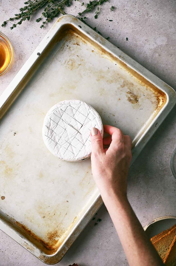 A hand places a round wheel of scored Baked Brie onto a baking sheet, with herbs and ingredients visible nearby.