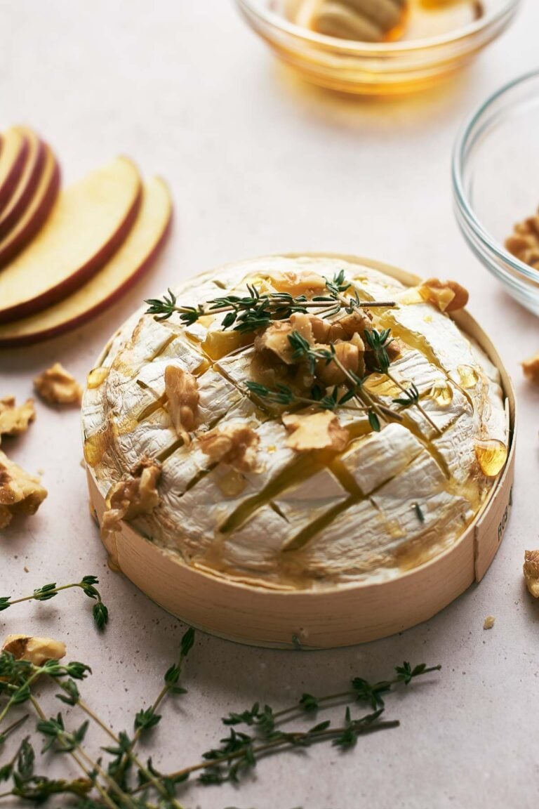 Round wheel of Baked Brie topped with walnuts, honey, and fresh thyme, served in a wooden container with apple slices and extra walnuts nearby.