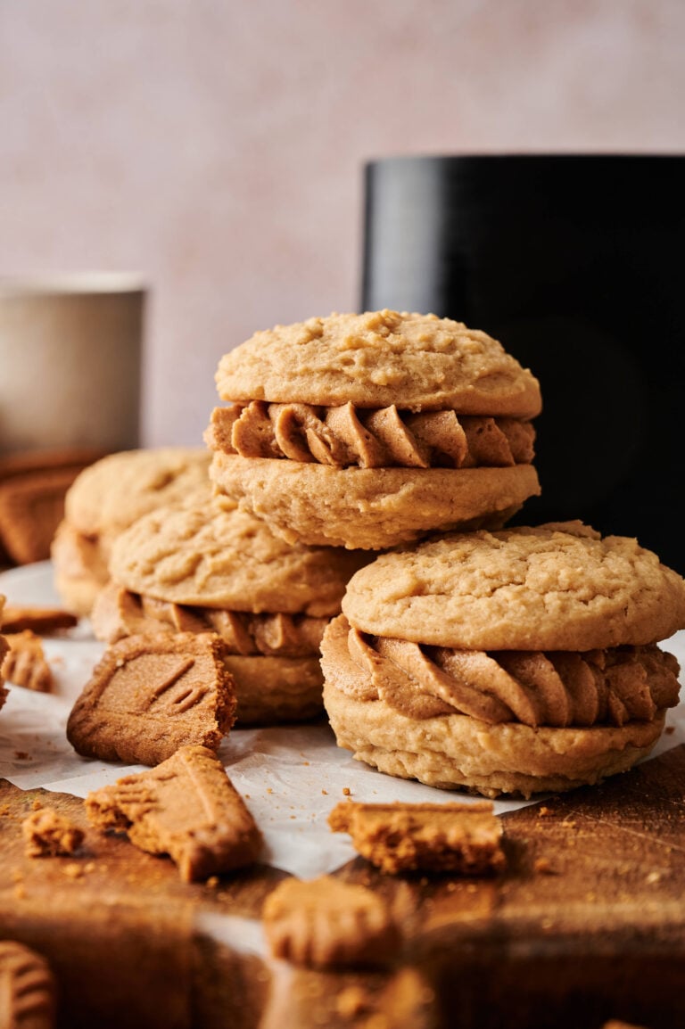 Three Air Fryer Biscoff Sandwich Cookies with creamy filling are stacked on parchment paper, surrounded by cookie crumbs and pieces, with a cup and a dark object in the background.