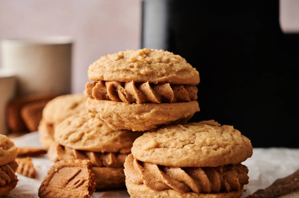Two oatmeal sandwich cookies filled with a thick layer of spiced cream, inspired by Air Fryer Biscoff Sandwich Cookies, with more cookies and a mug in the blurred background.