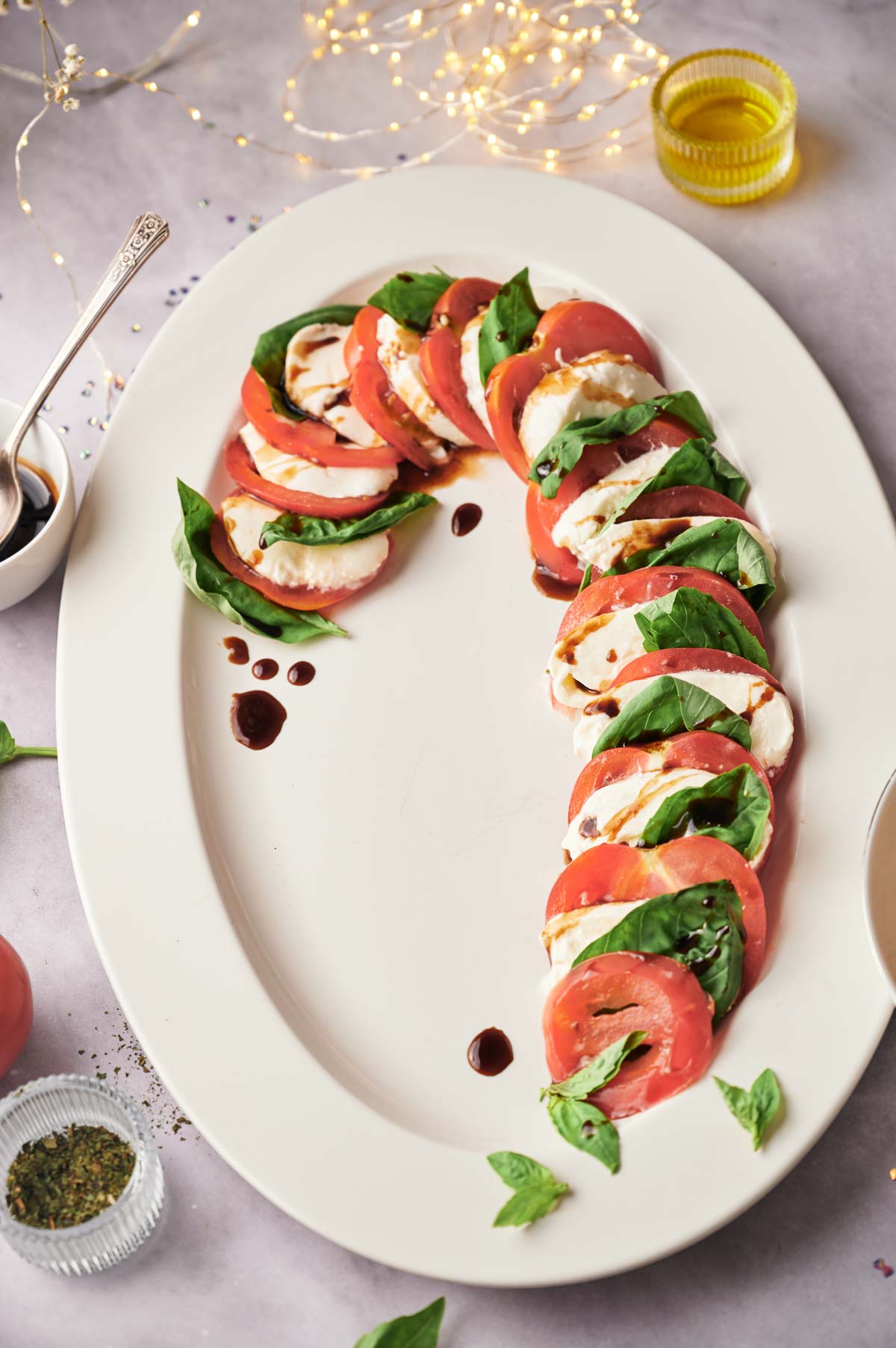 Candy Cane Caprese Board featuring sliced tomatoes, mozzarella, and basil arranged in a candy cane shape on a white platter, drizzled with balsamic glaze. Olive oil, spices, and festive lights in the background complete the look.