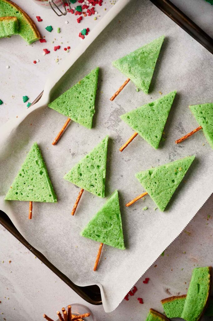 Green triangle-shaped Christmas Tree Sugar Cookies with pretzel sticks as trunks are arranged on a parchment-lined baking sheet, perfectly resembling festive Christmas trees.