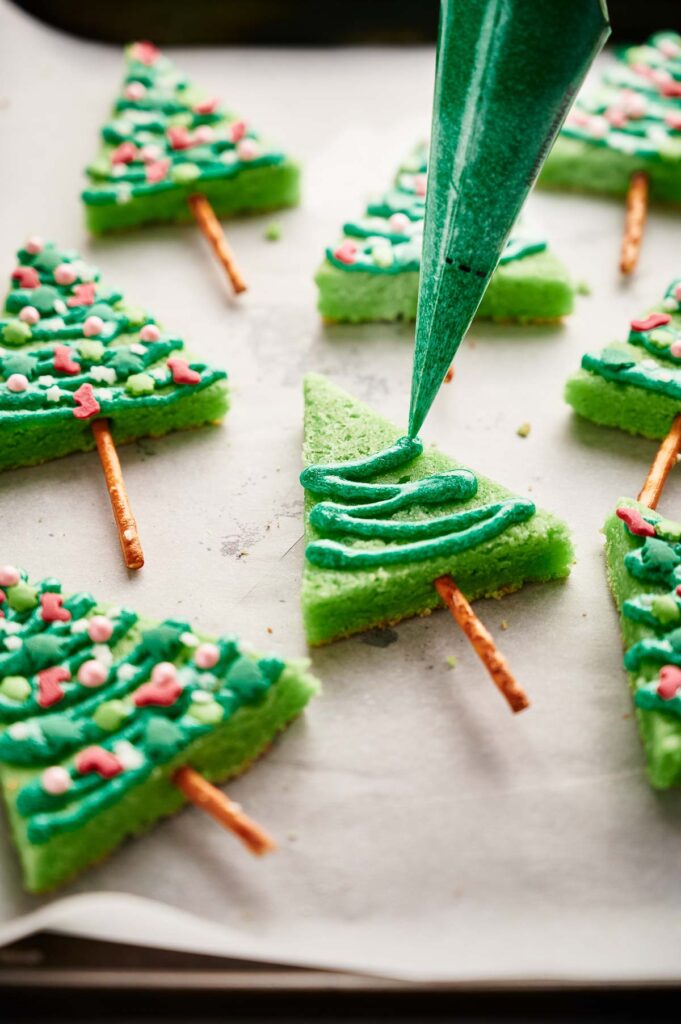 Rectangular Christmas Tree Sugar Cookies are being decorated with green icing from a piping bag. Each has a pretzel stick trunk and colorful sprinkles as ornaments, making them festive and fun to create.