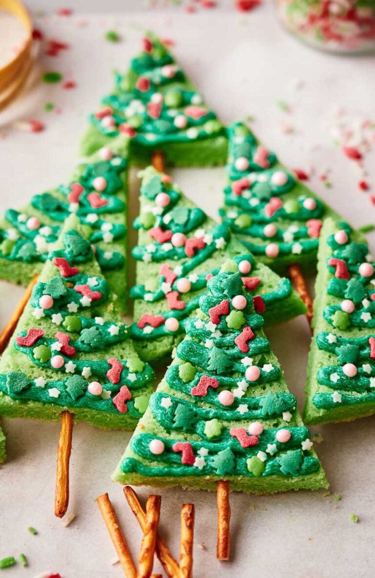 Green Christmas tree sugar cookies with frosting and sprinkles, each featuring a pretzel stick as the trunk, are artfully arranged on a light surface.