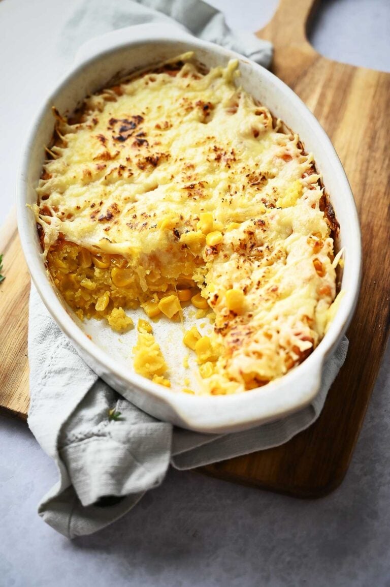 A baked Corn Casserole dish filled with cheesy corn pudding, with a portion scooped out, sits on a wooden board with a cloth napkin underneath.