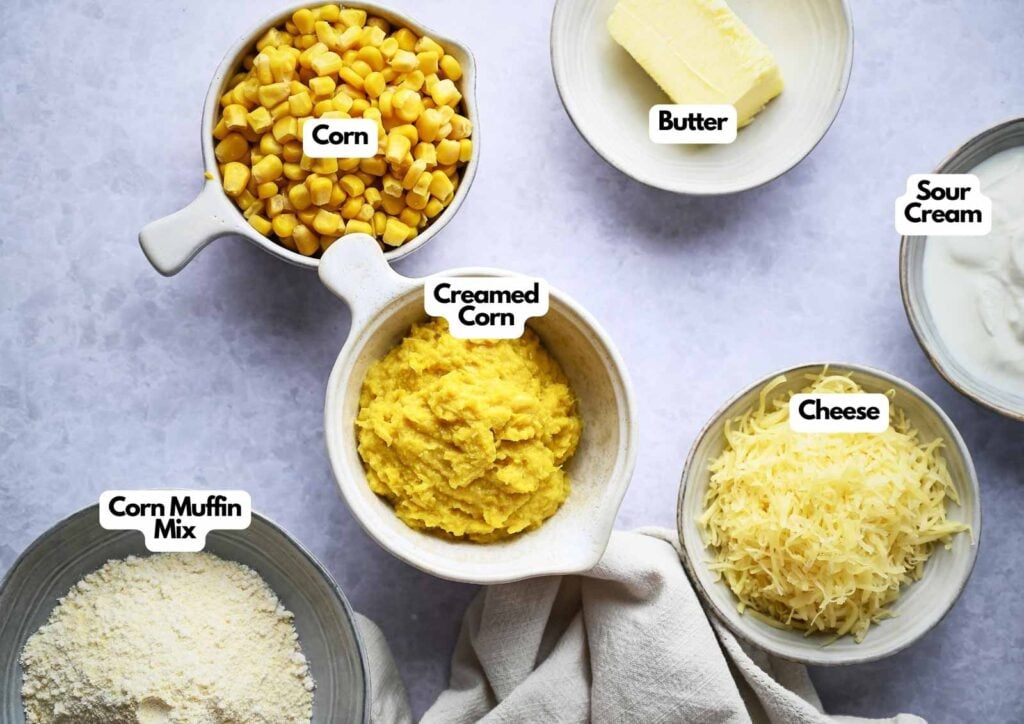 A flat lay of labeled ingredients in bowls for Corn Casserole: corn, creamed corn, butter, sour cream, shredded cheese, and corn muffin mix on a white surface.