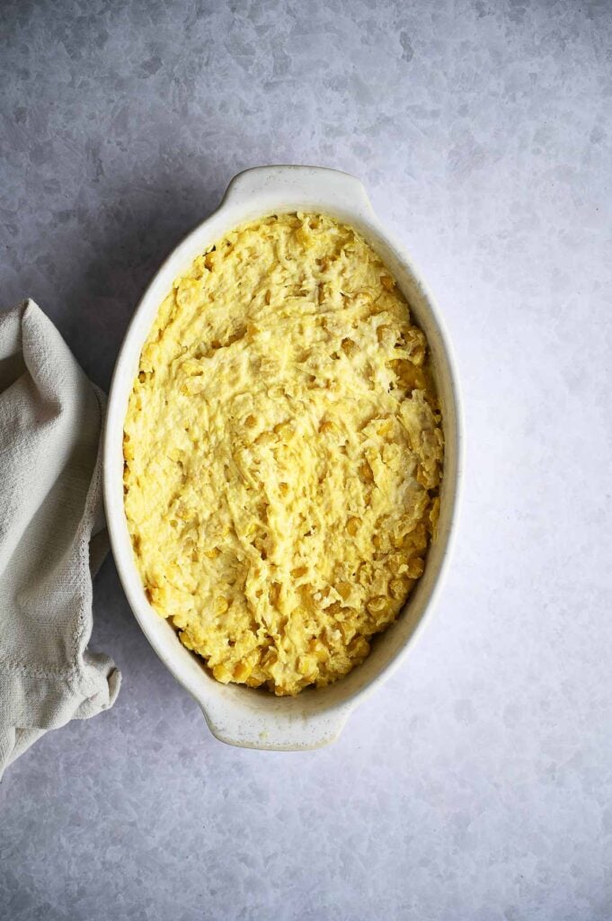 Oval white casserole dish filled with a yellow, textured corn casserole mixture, possibly baked, on a light gray surface next to a beige cloth.