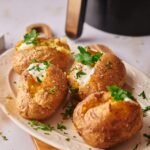 Four Air Fryer Baked Potatoes on a white plate, topped with sour cream and garnished with chopped parsley, sit on a table.