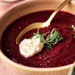 A bowl of Air Fryer Beet Soup topped with a dollop of cream, a sprig of rosemary, and a sprinkle of black pepper, with a spoon resting in the soup.