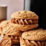 Three Air Fryer Biscoff Sandwich Cookies with a thick layer of brown cream filling are stacked on a plate, with more cookies and a cup blurred in the background.