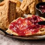 A plate of Air Fryer Scones, one split open and spread with red fruit jam, with a small bowl of jam and more scones in the background.