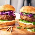 Two Air Fryer Veggie Burger Sliders with lettuce, sliced cucumber, purple cabbage, and sauce on a wooden board, each secured with a toothpick.