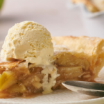A slice of Apple Pie topped with a scoop of vanilla ice cream on a white plate, with a spoon nearby.