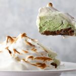 A slice of classic Baked Alaska with toasted meringue, revealing layers of green ice cream and cake on a white plate.