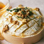 A round of Baked Brie cheese topped with walnuts, fresh thyme, and drizzled honey, presented in a wooden container with a bowl of honey beside it.