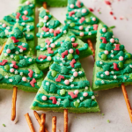 Green Christmas Tree Sugar Cookies decorated with icing and sprinkles, each with a pretzel stick trunk, are arranged on a white surface.