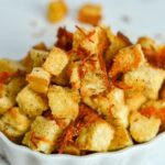 A bowl filled with golden-brown cheese croutons, perfectly seasoned and topped with extra grated cheese, sits on a white surface.