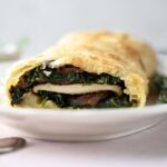 A close-up of a Mushroom Wellington pastry roll, sliced to reveal layers of spinach, mushrooms, and caramelized onions on a white plate.