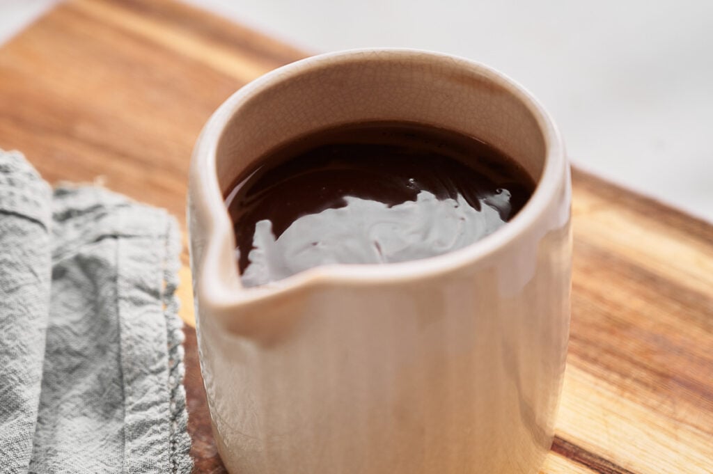 A small ceramic pitcher filled with rich hot fudge sauce sits on a wooden surface next to a folded gray cloth.