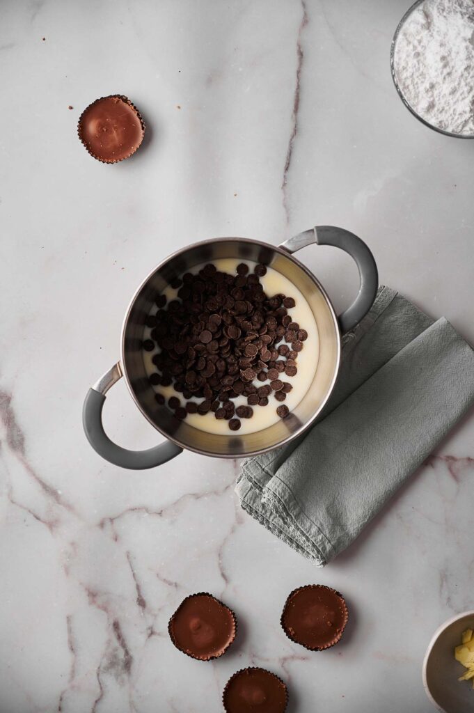 A saucepan filled with chocolate chips and condensed milk sits on a gray cloth, ready to become rich Hot Fudge Sauce, surrounded by peanut butter cups and baking ingredients on a marble surface.