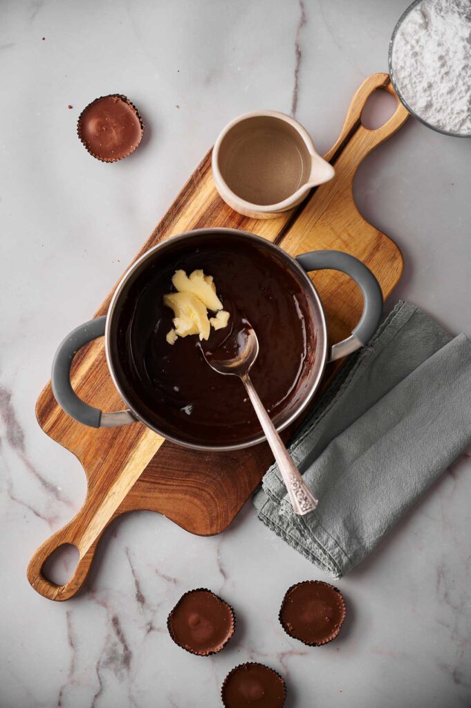 A pot of melted chocolate with hot fudge sauce and butter sits on a wooden board next to a small jug, gray napkin, and several empty chocolate molds.