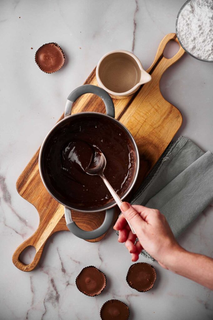 A hand stirs melted chocolate for hot fudge sauce in a saucepan on a wooden board, surrounded by empty cupcake liners, a cup of liquid, and a bowl of powdered sugar.