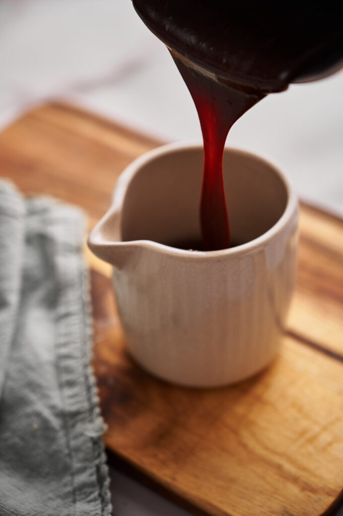 Hot Fudge Sauce being poured from a container into a small white ceramic pitcher on a wooden surface, next to a folded gray cloth.