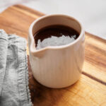 A small ceramic pitcher filled with rich hot fudge sauce sits on a wooden board next to a folded gray cloth.