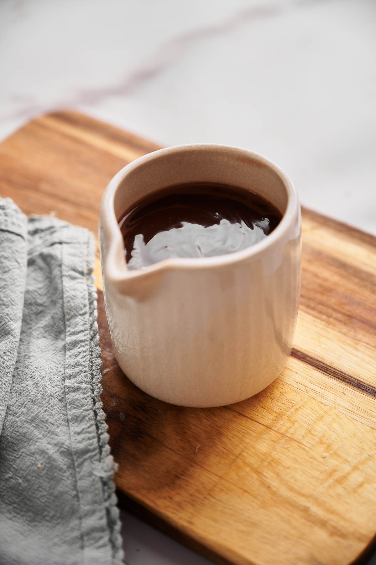 A small ceramic pitcher filled with rich hot fudge sauce sits on a wooden board next to a folded gray cloth.