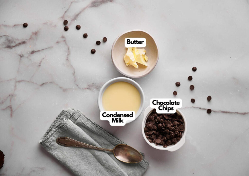A marble surface with bowls of butter, condensed milk, and chocolate chips—ingredients for Hot Fudge Sauce—plus a spoon, folded napkin, and scattered chocolate chips.