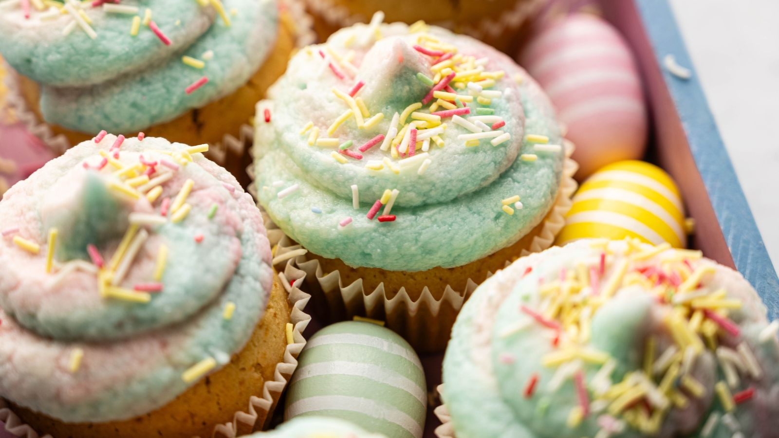 Cupcakes with pastel blue and pink frosting, topped with sprinkles, are arranged next to decorated pastel Easter eggs in a wooden tray.