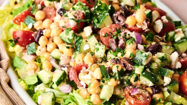 A close-up of a salad with chickpeas, cherry tomatoes, cucumber, red onion, kalamata olives, feta cheese, and chopped parsley on a bed of lettuce.