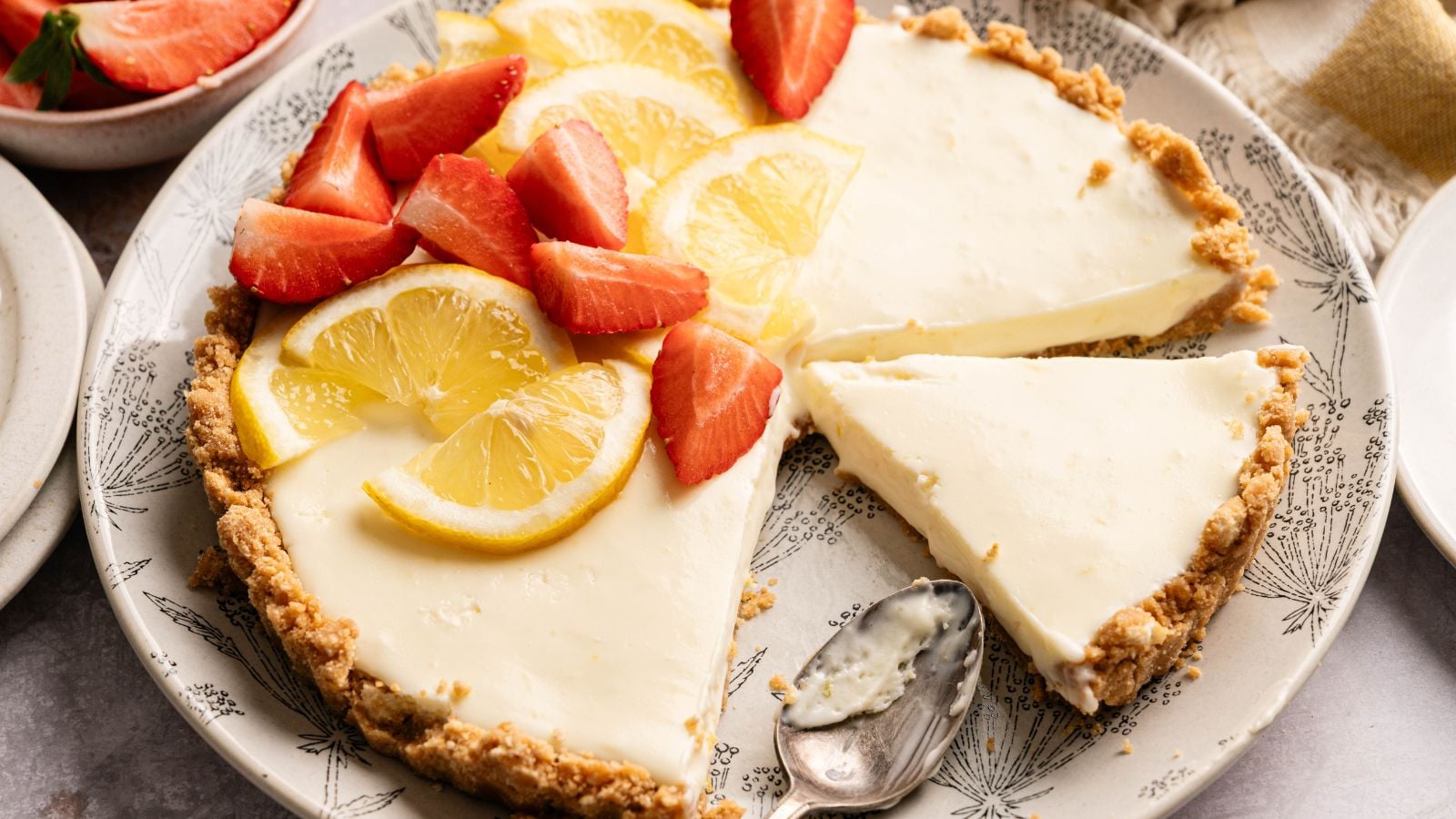 A creamy pie with a crumbly crust, topped with sliced lemons and strawberries. One slice has been removed and a spoon rests on the plate.
