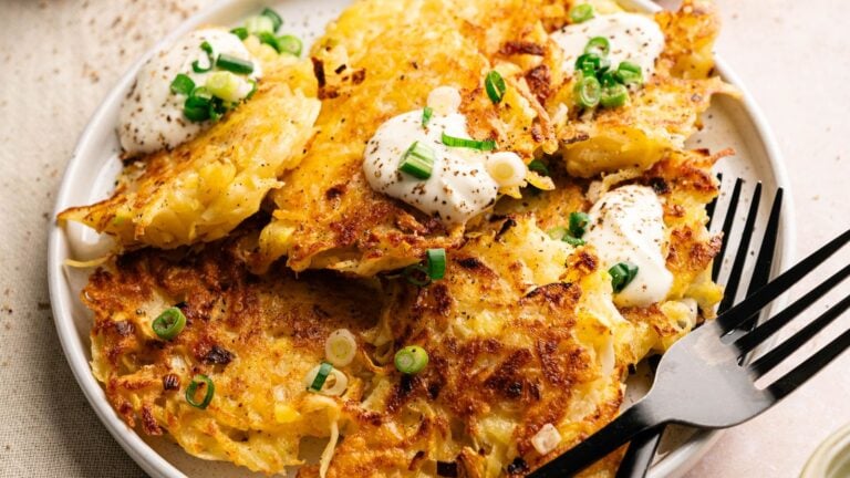 A plate of crispy potato latkes topped with sour cream and chopped green onions, with a fork resting on the side.
