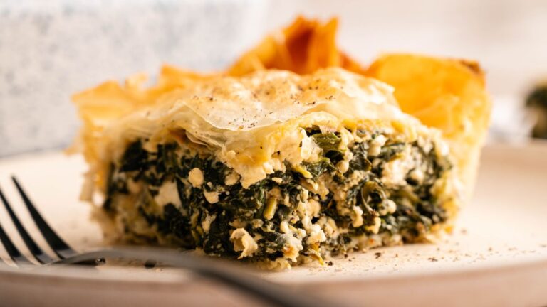 A close-up of a slice of spinach and cheese pie with a flaky golden crust on a plate, with a fork resting nearby.
