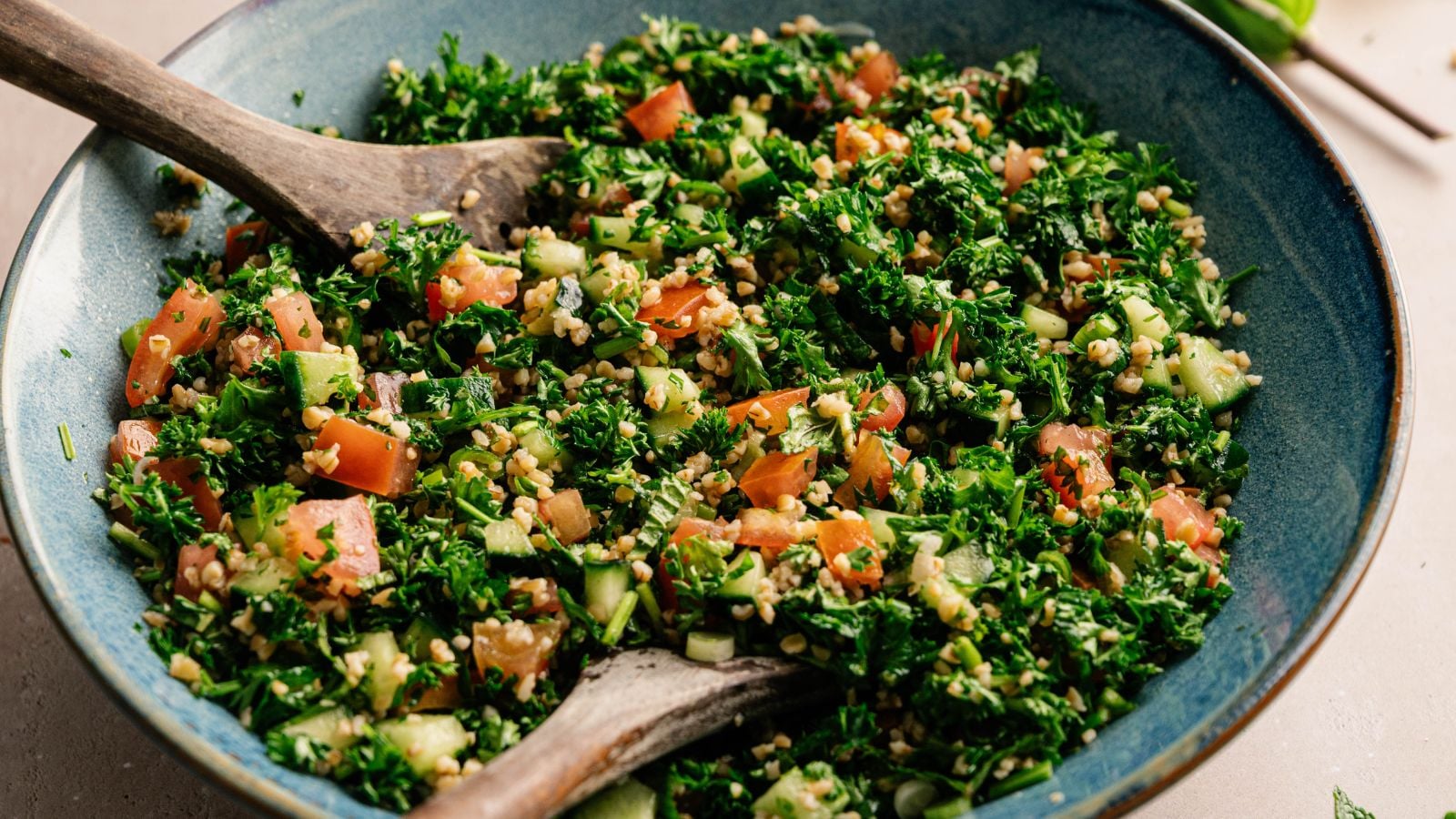 A bowl of tabbouleh salad with chopped parsley, tomatoes, cucumbers, and bulgur, served with wooden utensils.