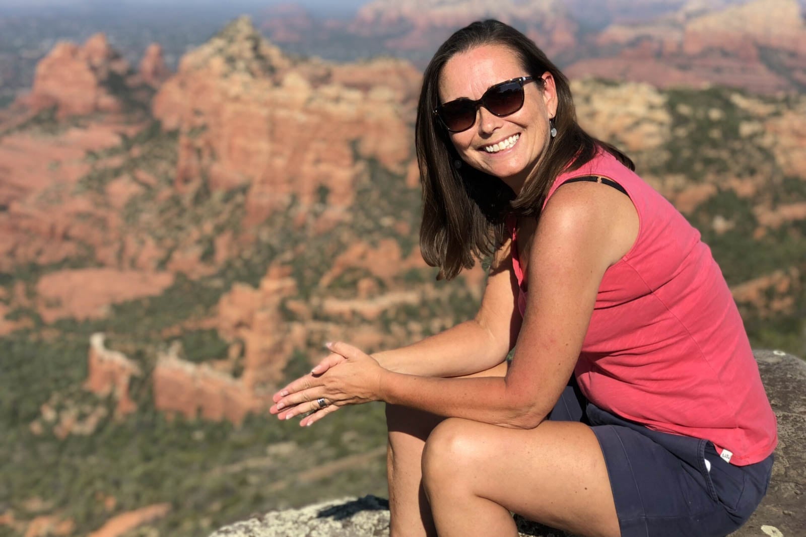 A woman wearing sunglasses and a pink sleeveless shirt sits on a rock, smiling, with a sunny desert landscape and red rock formations in the background.