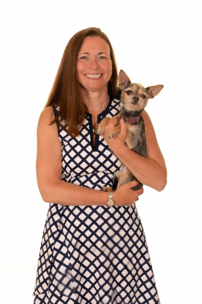 A woman in a sleeveless, patterned dress smiles while holding a small dog with pointy ears against a plain white background.