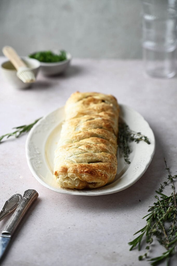 A braided puff pastry loaf, reminiscent of a classic Mushroom Wellington, is served on a white plate with sprigs of fresh herbs, next to cutlery and a bowl of chopped greens.