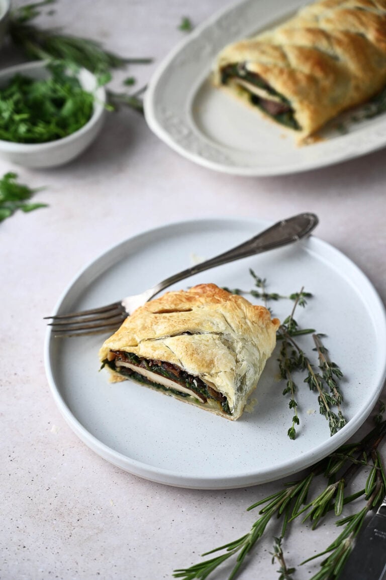 A slice of Mushroom Wellington puff pastry on a white plate with a fork, garnished with fresh herbs; the remaining pastry is on a platter in the background.