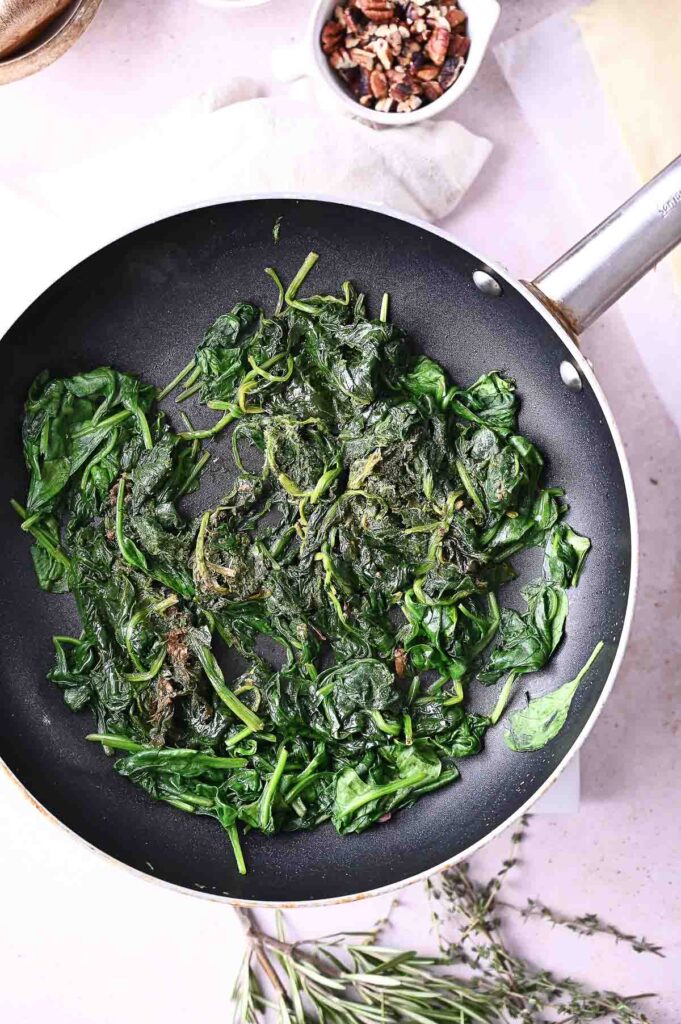 Sautéed spinach cooking in a black nonstick frying pan, with some herbs visible nearby and a small bowl of chopped nuts in the background—perfect ingredients for assembling a flavorful mushroom wellington.