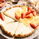A no bake lemon tart cheesecake with a graham cracker crust, garnished with sliced lemons and strawberries, sits on a patterned plate with one slice partially removed.