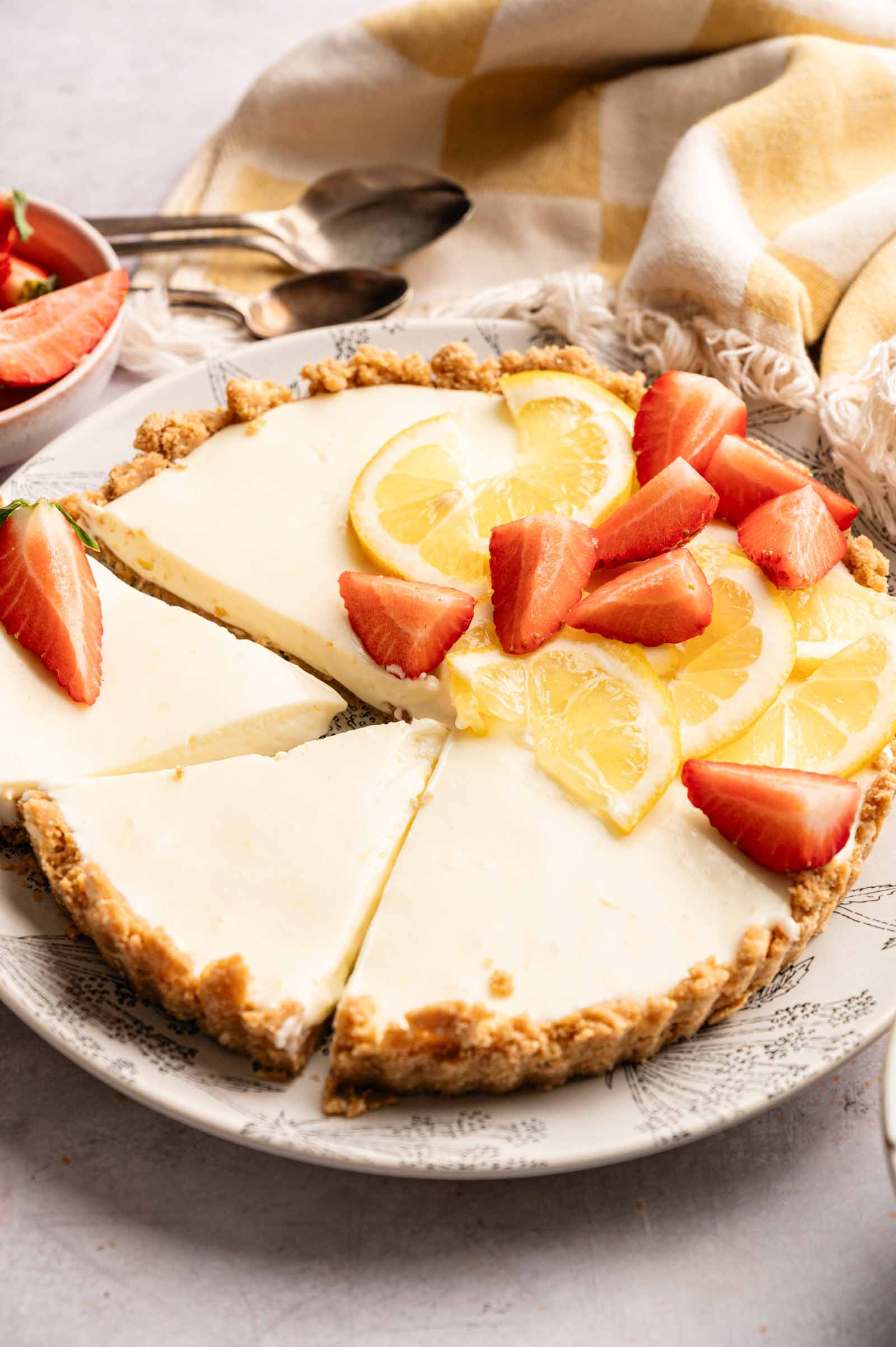 A no bake lemon tart cheesecake with a graham cracker crust, garnished with sliced lemons and strawberries, sits on a patterned plate with one slice partially removed.