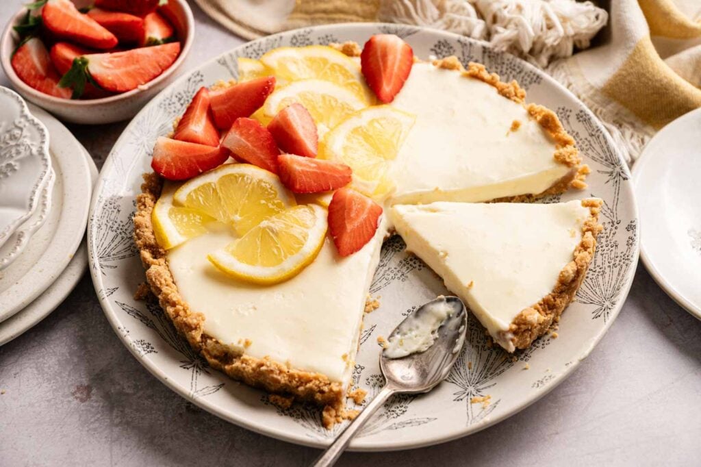 A creamy No Bake Lemon Tart with a crumb crust is topped with sliced lemons and strawberries. One slice is missing, and a spoon rests on the plate. Nearby are small plates and a bowl of strawberries.