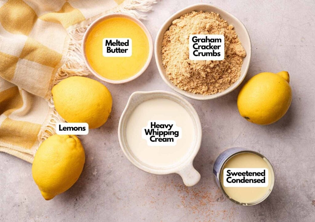 Top-down view of No Bake Lemon Tart ingredients on a counter: melted butter, graham cracker crumbs, lemons, heavy whipping cream, and sweetened condensed milk, each labeled.