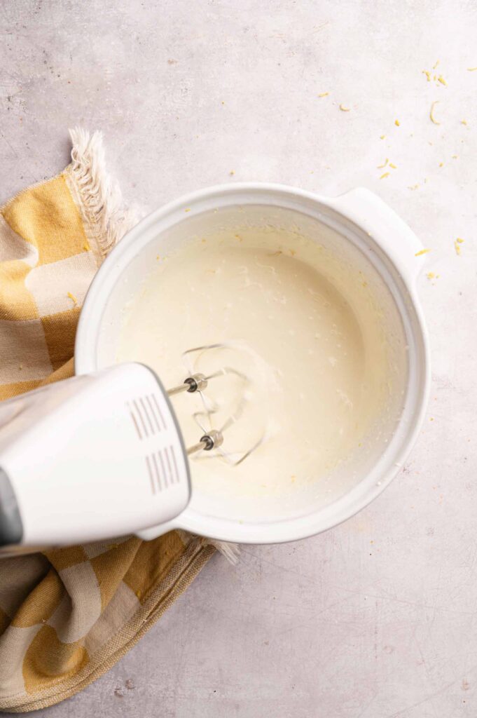 An electric hand mixer blending a creamy mixture for a No Bake Lemon Tart in a white bowl, placed on a light surface with a yellow and white checkered cloth nearby.