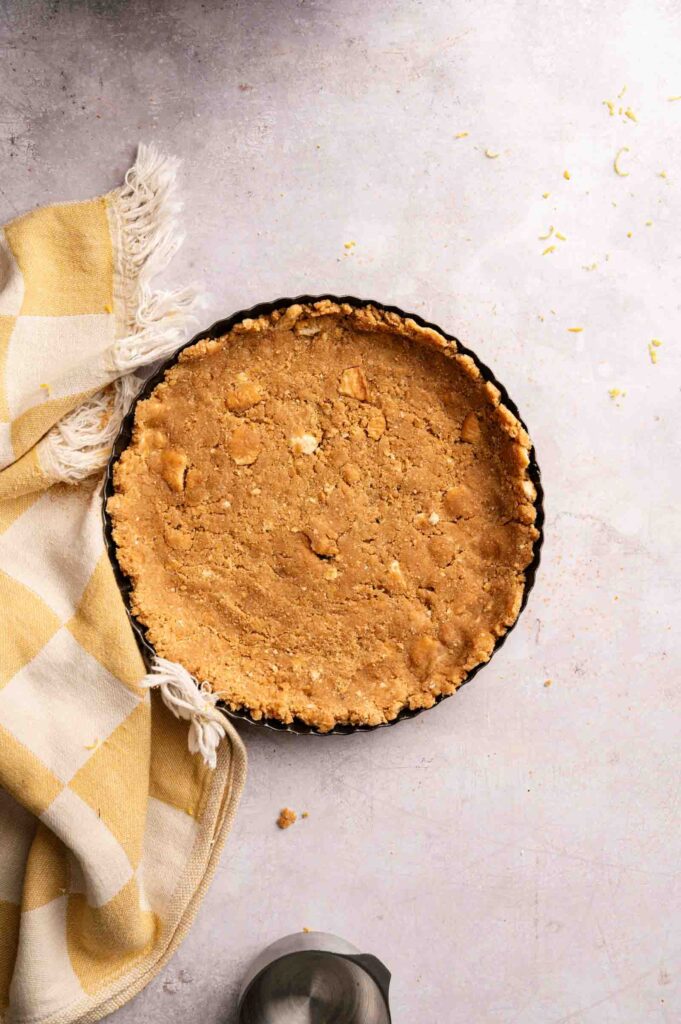 A round, unbaked pie crust for a No Bake Lemon Tart rests in a tart pan on a light surface beside a folded yellow and white checkered cloth.