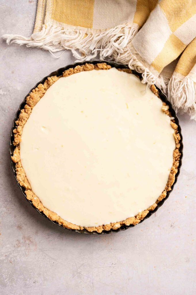 A round pie pan with a graham cracker crust and creamy white filling, this No Bake Lemon Tart sits on a light surface next to a yellow and white checkered cloth.